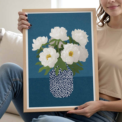 A person sitting on a couch holds up a framed art print featuring white peony flowers in a blue and white dotted vase against a dark blue background.