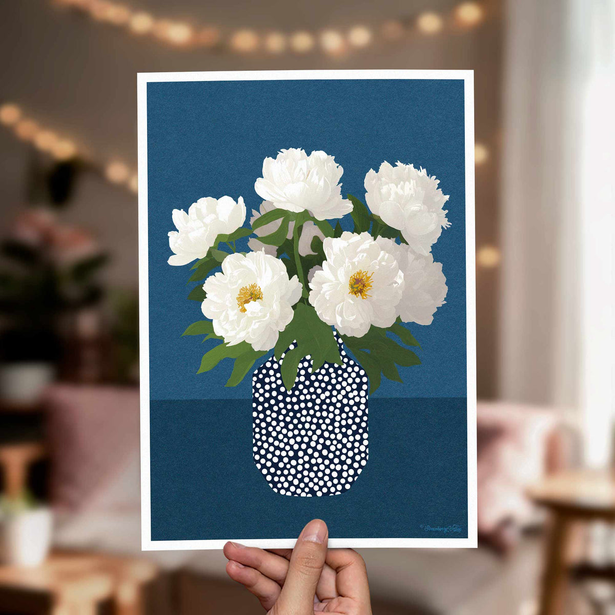 A hand holds a square art print featuring white peony flowers in a white vase with green leaves against a dark blue background. The print is shown in a softly blurred living room setting with warm string lights in the background.