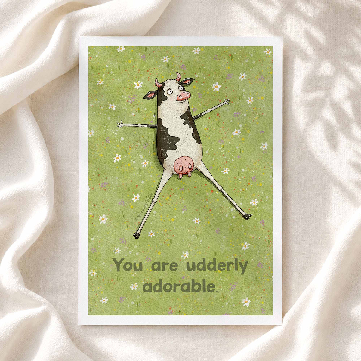 A square greeting card featuring a whimsical illustration of a black-and-white cow with arms and legs made of sticks, set against a light green floral background with the text 'You are udderly adorable.' The card is photographed on draped white fabric.